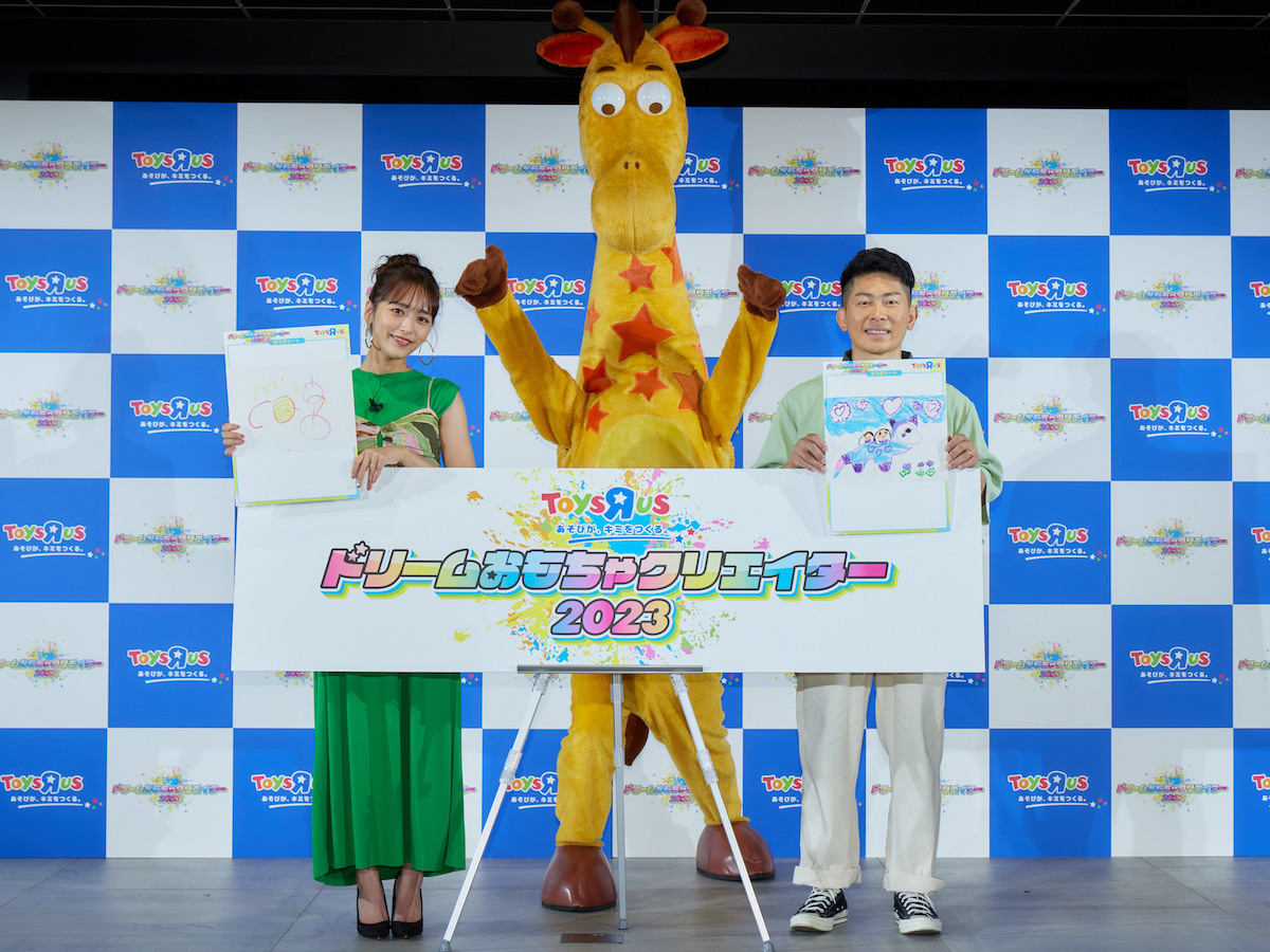 トイザらスの公式キャラクター キリンの「ジェフリー」を迎えて写真撮影に応じた近藤千尋と太田博久。手には娘たちが描いた絵を持っている